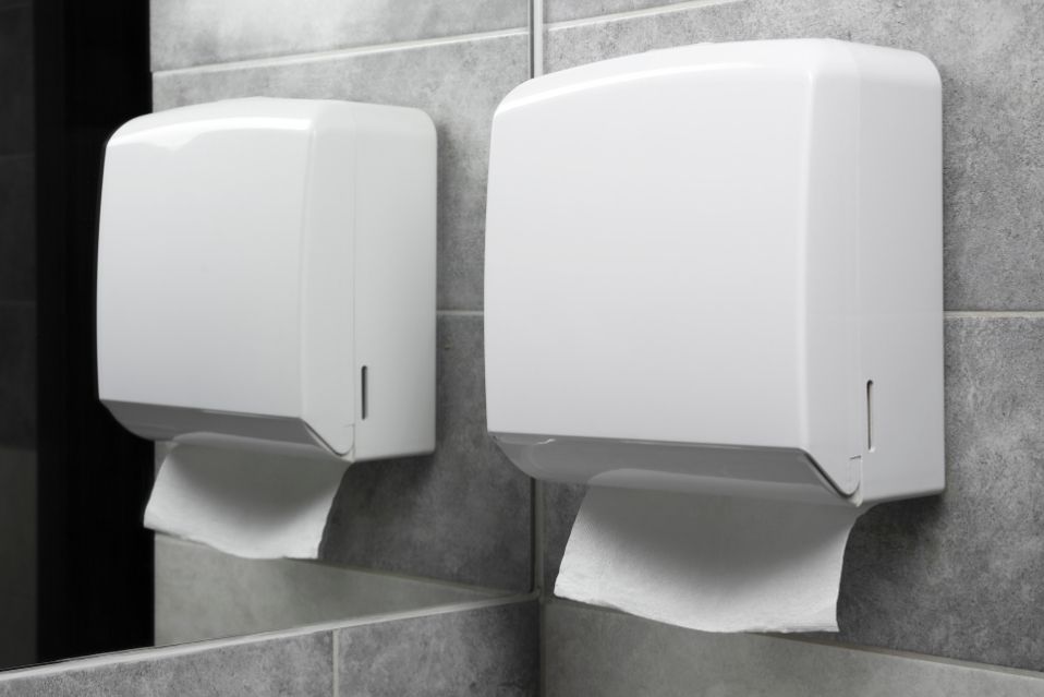 Um dispenser de papel toalha de rolo, colocado em um ambiente interno, em frente a um espelho.