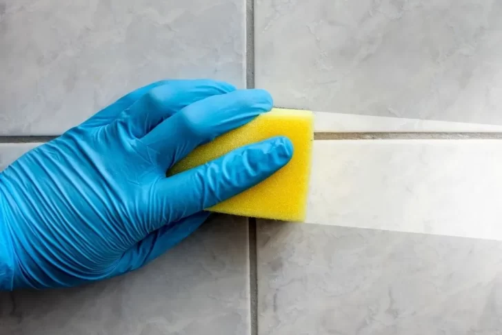Mão com luva azul limpando azulejo com esponja amarela, mostrando o antes e depois da limpeza.
