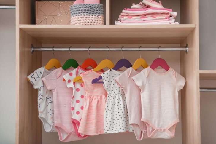 guarda-roupa de bebê com bodies e vestidinhos em cabides coloridos; prateleira superior com roupas dobradas, cesta de tricô e caixa organizadora.