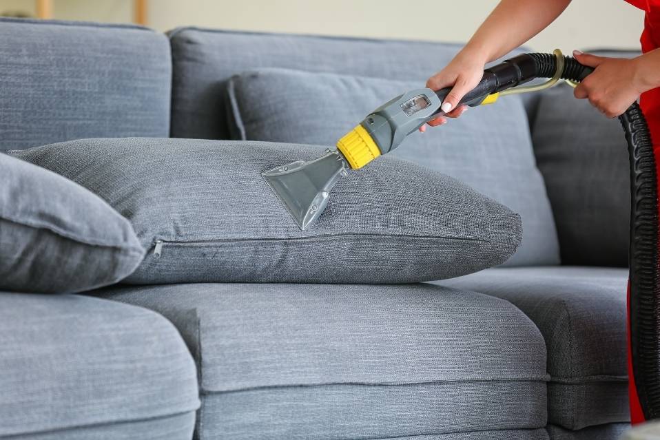 mãos usando extratora/aspirador em almofada de sofá cinza, realizando limpeza a seco; estofado cinza ao fundo.