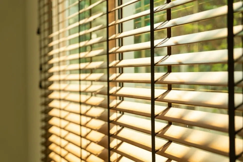 persiana horizontal de lâminas de madeira semiaberta, com luz do sol atravessando e fundo verde desfocado.