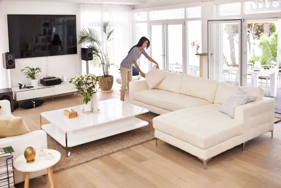 Uma mulher em uma sala de estar moderna e clean arruma as almofadas de um grande sofá branco, com o objetivo de manter a casa organizada. A sala tem paredes brancas, piso de madeira e grandes portas de vidro que dão para um pátio. A iluminação é natural e a decoração é minimalista, com vasos de plantas e um rack de TV com poucos eletrônicos.
