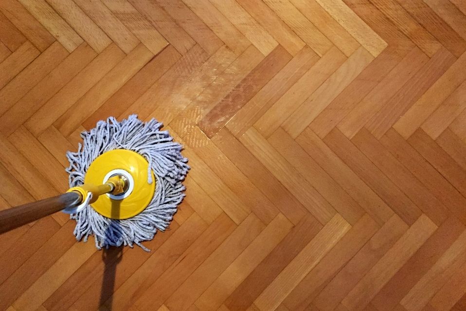Um esfregão está sendo usado para limpar madeira em um piso com padrão espinha de peixe. A foto aérea mostra o esfregão molhado sobre a superfície do chão de madeira, destacando o processo de limpeza.