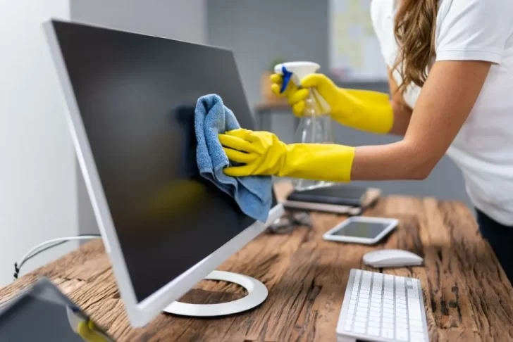 Pessoa usando luvas amarelas, limpando um monitor de computador com um pano azul e um produto de limpeza em spray. A cena mostra o ambiente de trabalho e enfatiza a higienização de eletrônicos.