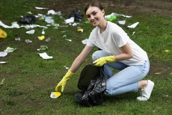 Jovem com luvas amarelas, agachada em um gramado, sorrindo e colocando lixo em um saco preto. A grama ao redor está suja com diversos resíduos, como copos plásticos e sacolas, destacando a ação de limpeza e a preocupação com o meio ambiente.
