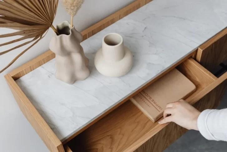 Visão de perto de uma cômoda de madeira clara, com um tampo de mármore branco embutido. Sobre o mármore, há dois vasos de cerâmica de formatos orgânicos e uma folha seca de palmeira. Uma mão está colocando um objeto em uma das gavetas abertas.