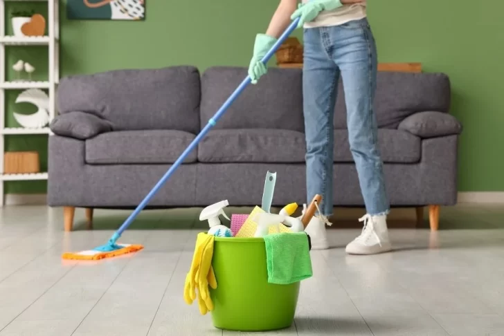 Uma pessoa com calças jeans e luvas de limpeza esfrega o chão de uma sala de estar com um esfregão. Em primeiro plano, um balde verde brilhante está cheio de produtos de limpeza, esponjas e luvas amarelas. O ambiente tem uma parede verde escura e um sofá cinza.